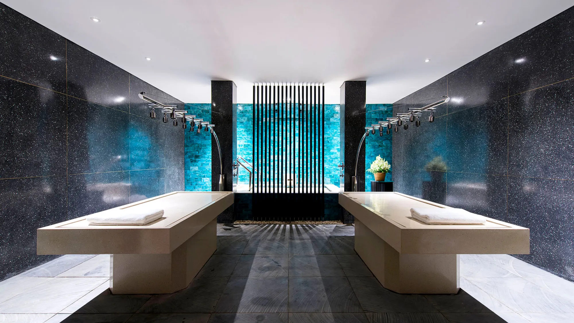 A sleek, symmetrical spa treatment room featuring two light-colored stone massage tables. Above each table hangs a metal, multi-nozzle Vichy shower fixture. The room is decorated with dark, speckled stone walls on the sides and vibrant, glowing turquoise blue tiles on the back wall, which is partially obscured by a central vertical black slatted partition. The floor is made of large, dark grey tiles, and a rolled white towel rests at the end of each table.