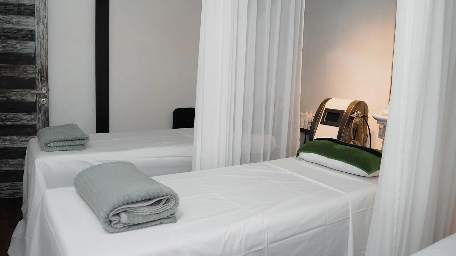 A bright, clean spa treatment room featuring two massage beds dressed in crisp white linens. The bed in the foreground has a neatly folded light grey towel at the foot and a dark green cushion at the head. Sheer white curtains hang from the ceiling to provide privacy between the stations. In the background to the right, a modern cosmetic skincare machine with a digital screen is visible next to some product bottles, while a dark, rustic wooden door stands on the left.