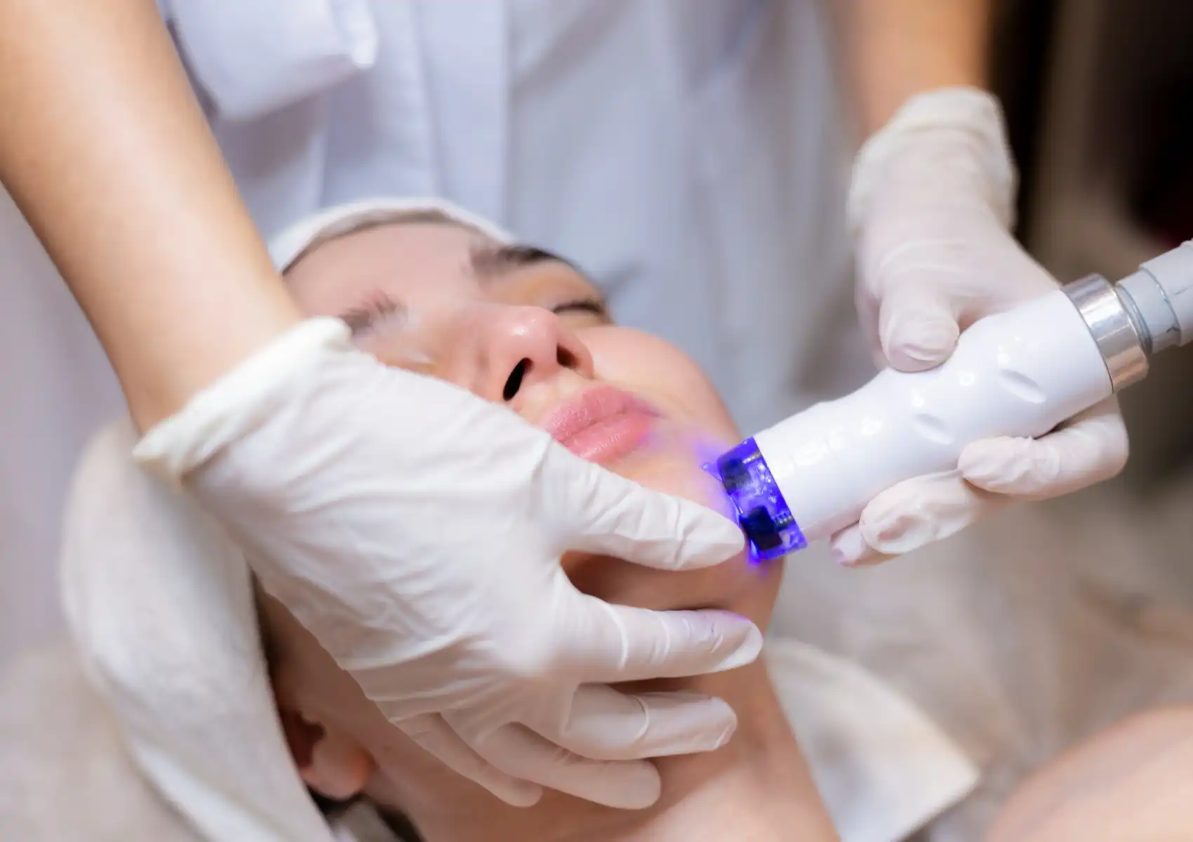 infuse-ultrasound A gloved esthetician using a white handheld ultrasound or LED facial device emitting blue light on a woman's lower face and lip area during a clinical skin treatment