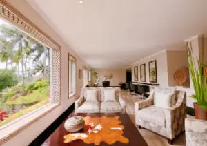 Elegant spa lounge with patterned armchairs, a wooden coffee table, and salon chairs, overlooking a lush tropical garden with palm trees through a large window.