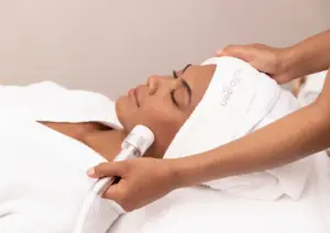 A woman lying down with eyes closed wearing a white Pollogen headband receiving an OxyGeneo facial treatment, with a therapist applying a white handheld device to her cheek