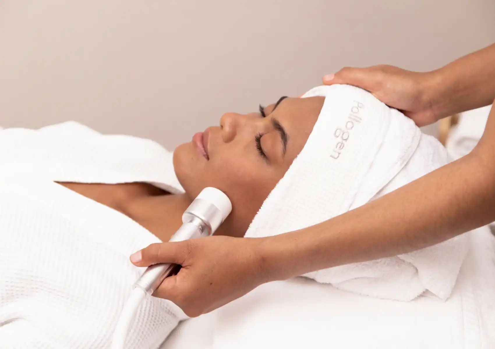 A woman lying down with eyes closed wearing a white Pollogen headband receiving an OxyGeneo facial treatment, with a therapist applying a white handheld device to her cheek