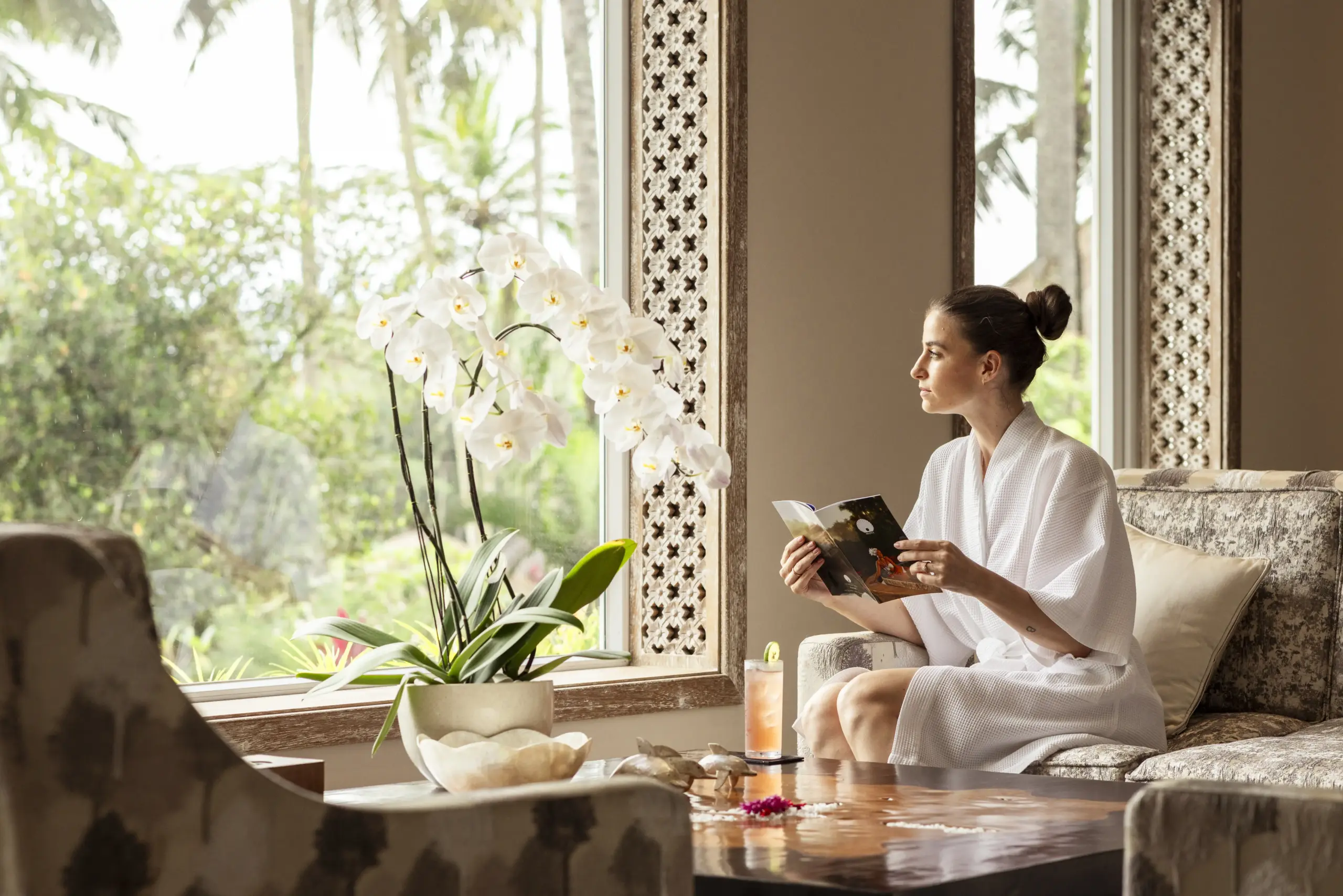 spa-with-valley-of-the-king-view A woman in a white spa robe sits on an ornate sofa reading a menu or brochure, surrounded by white orchids, a tropical drink, and carved Balinese window frames overlooking lush palm trees.