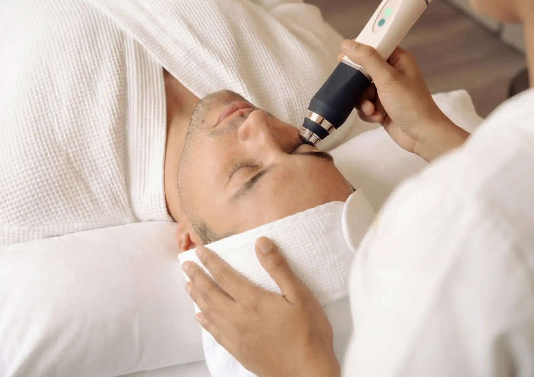 super-facial-akoya Man receiving a facial treatment with a handheld skincare device applied near his nose, lying back on a spa bed wrapped in white towels.