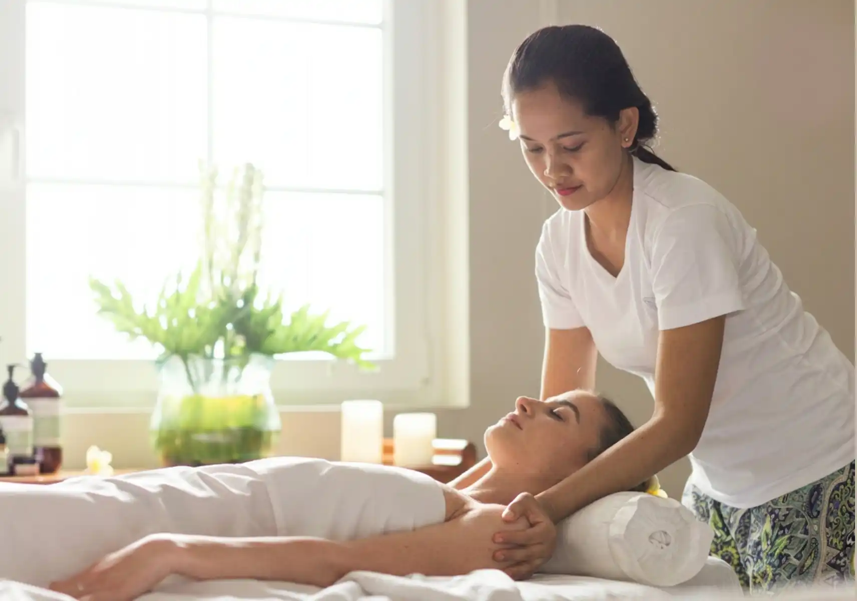traditional-balinese-massage-spa-bali A Balinese spa therapist wearing a white uniform and frangipani flower in her hair performs a traditional massage on a woman lying on a treatment table, with candles and tropical plants visible in the background.