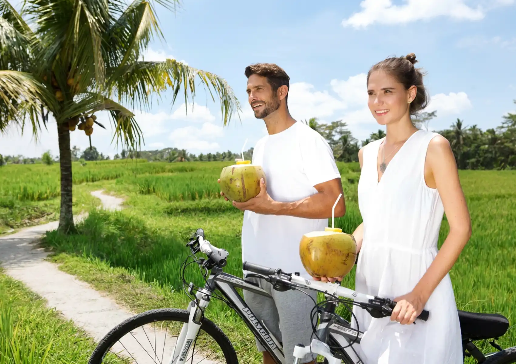 wellness-retreat-bali Cyclists enjoying fresh coconut water at a Bali spa retreat, standing in a tropical field with palm trees and blue sky.