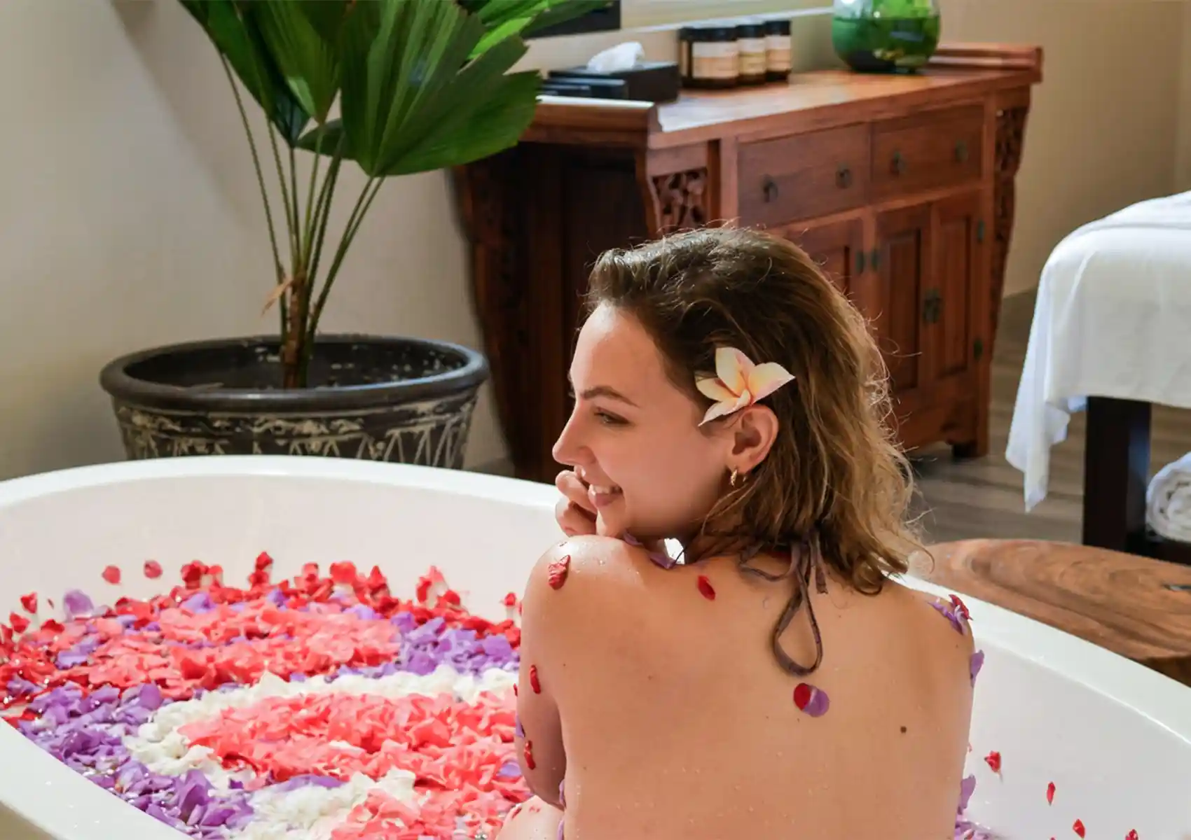 balinese-flower-bath A smiling woman sitting in a white bathtub filled with a vibrant pattern of red, pink, white, and purple flower petals, relaxing after a rejuvenating body scrub Bali treatment.