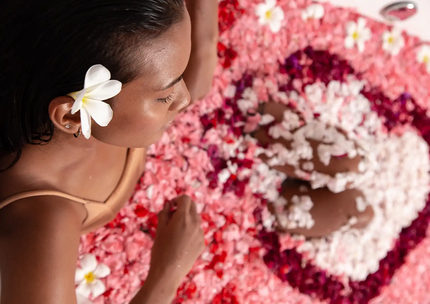 balinese-flower-bath A serene woman with a white frangipani flower tucked behind her ear soaks in a luxurious bath thickly covered in a vibrant pattern of pink, red, and white petals during a relaxing wellness treatment Ubud.