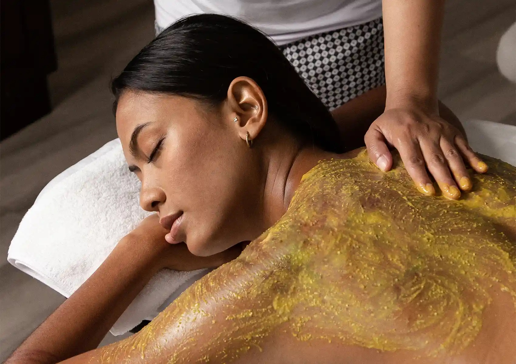 A relaxed woman lying face down on a white towel while a spa therapist massages a yellow, textured herbal paste onto her back during a traditional body scrub Bali treatment.