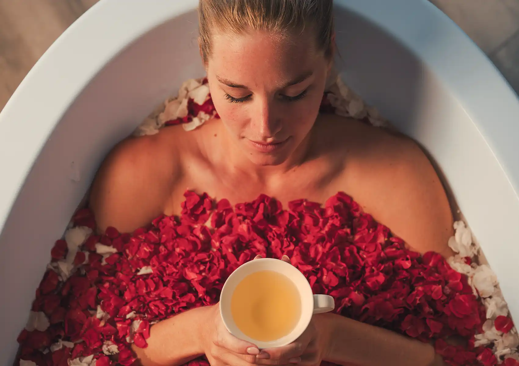 A tranquil woman submerged in a white bathtub filled with rich red rose petals holds a delicate cup of tea, enjoying a peaceful wellness treatment Ubud.