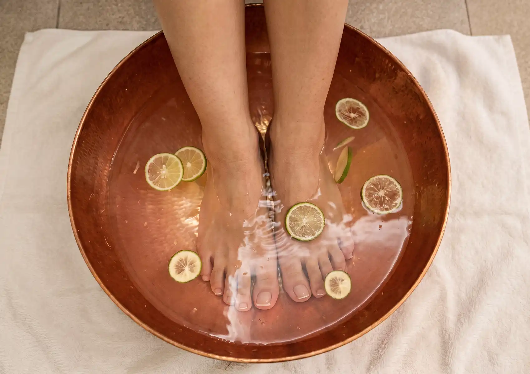 foot-bath-arrival An overhead shot of a person's bare feet soaking in a large round copper bowl filled with clear water and floating slices of fresh lime, resting on a white towel as a refreshing introduction to reflexology ubud.