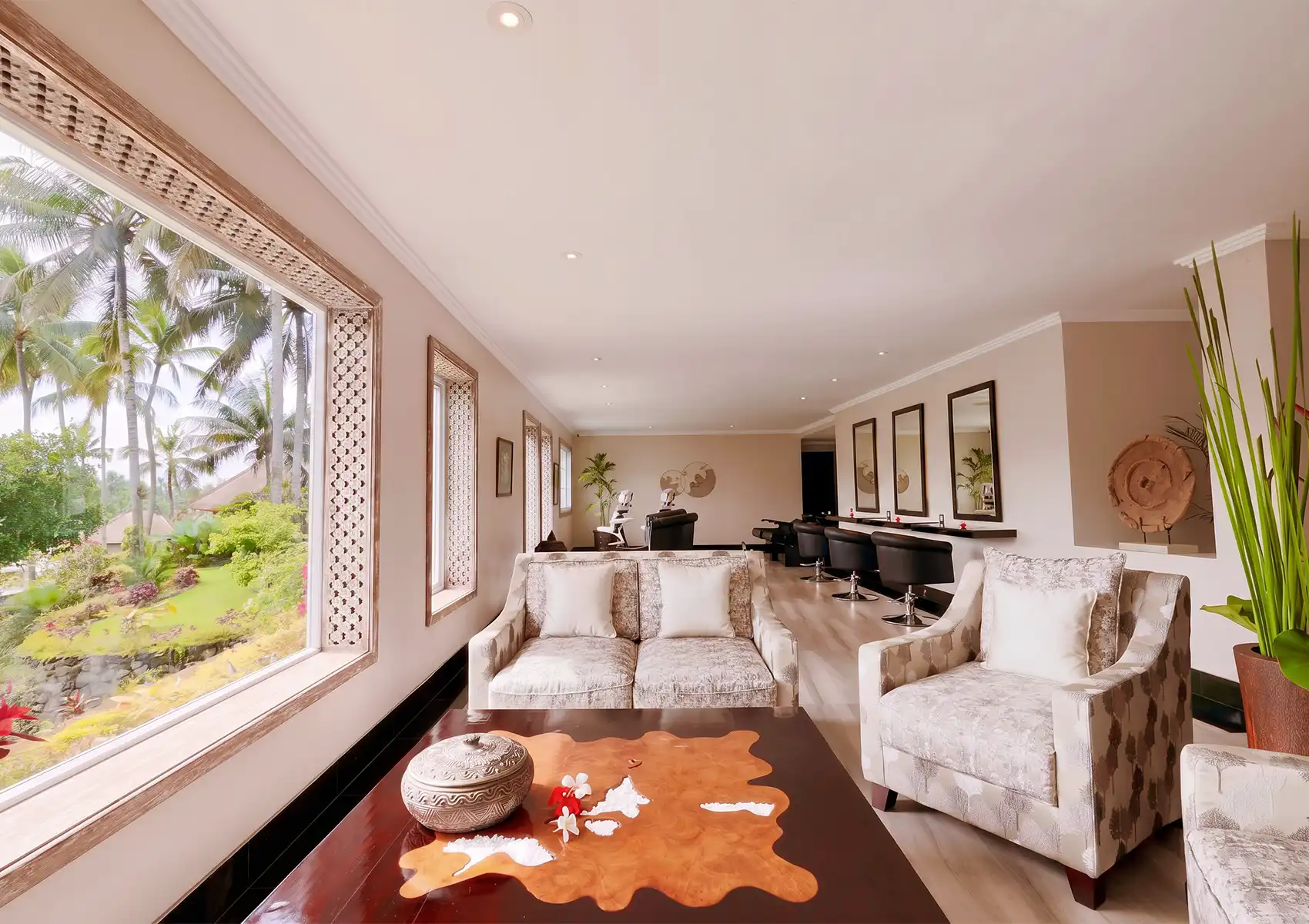 interior-akoya-spa-bali The spacious, airy interior lounge of a spa featuring patterned armchairs, a decorative wooden coffee table, and large picture windows framing a vibrant tropical garden landscape, creating a tranquil environment for reflexology ubud.