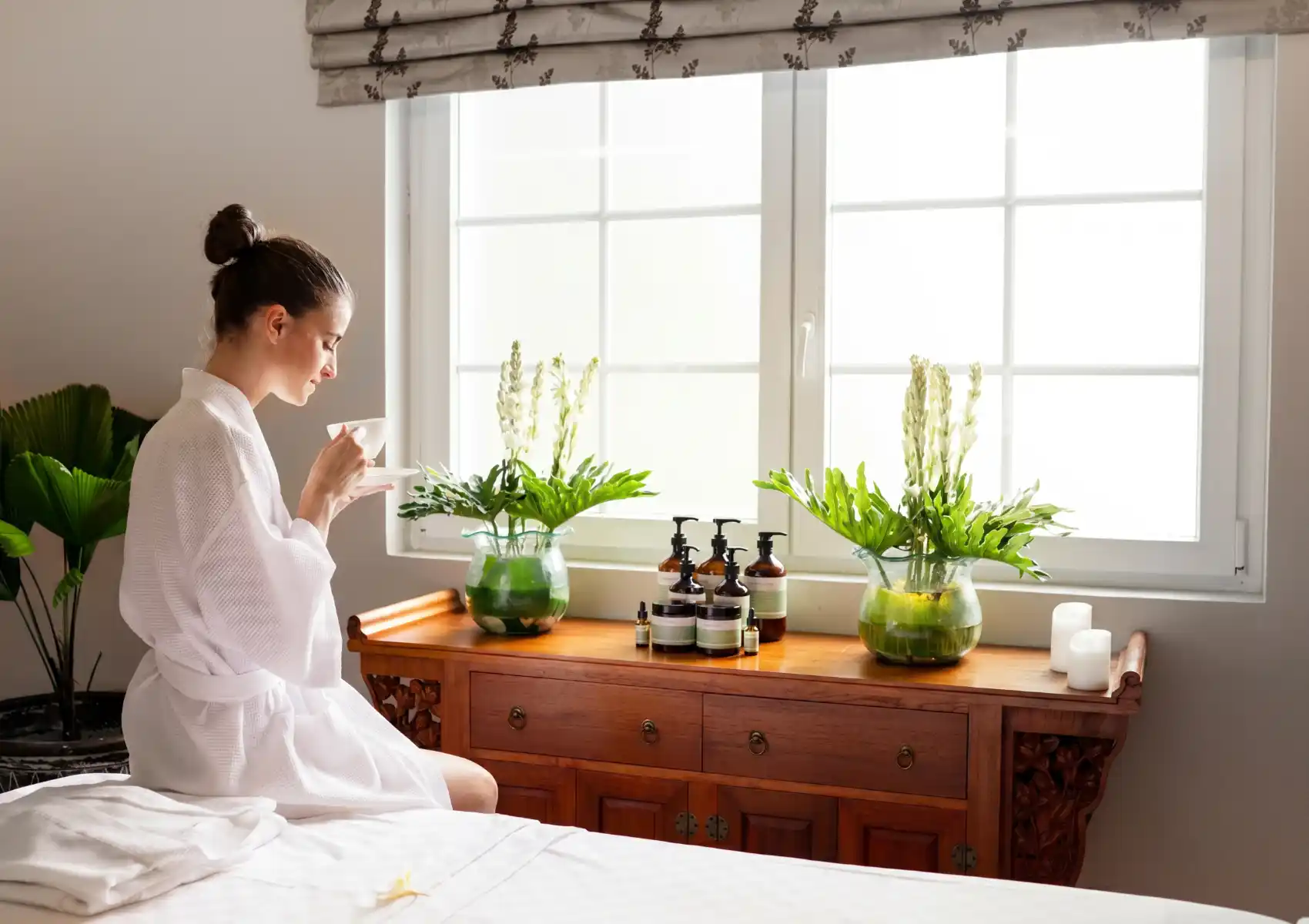 luxury-spa-day-ubud A woman in a white bathrobe peacefully sips tea by a bright window at a luxury spa in Ubud, sitting next to a wooden console table displaying skincare products and fresh floral arrangements.
