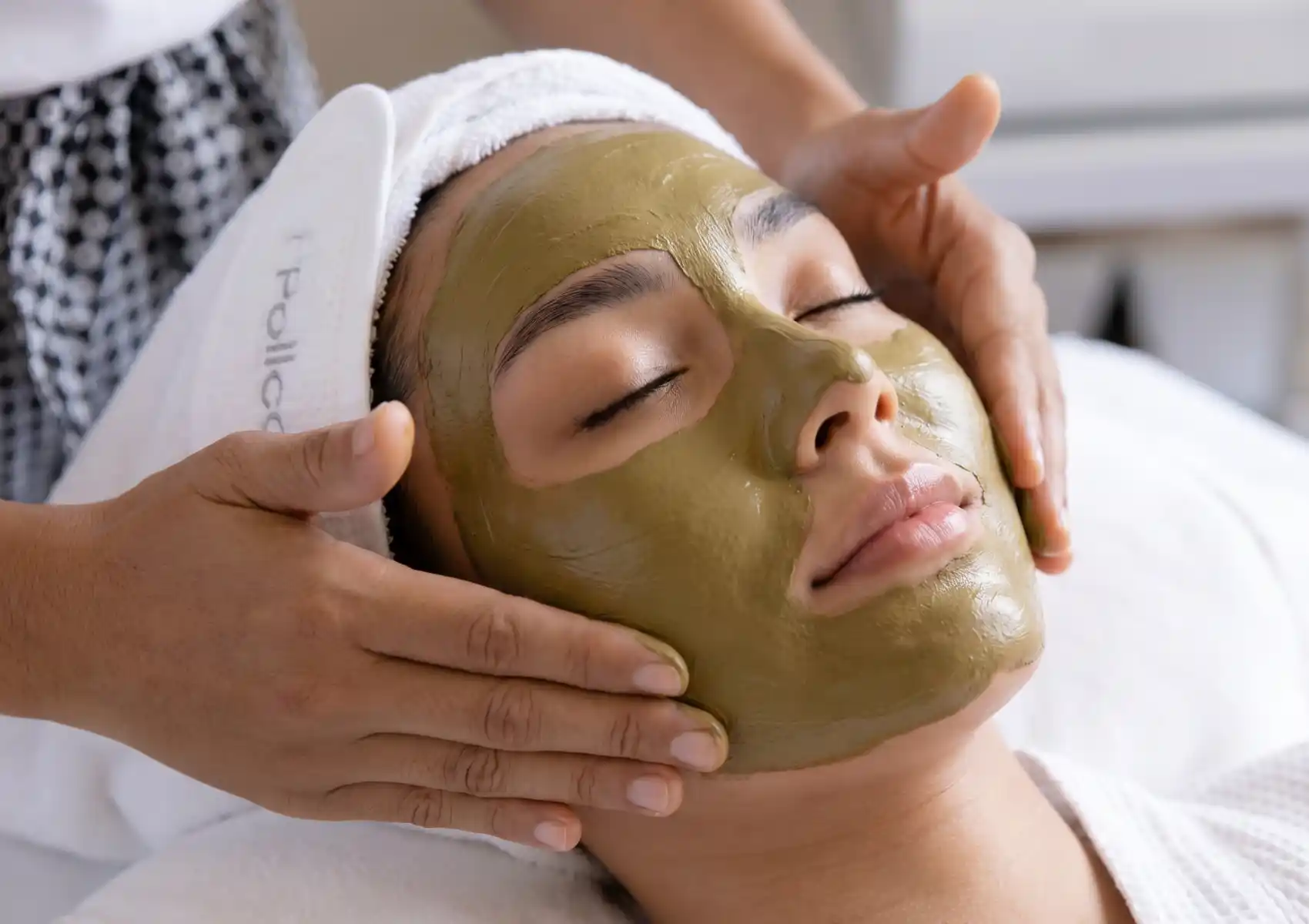A relaxed woman with her eyes closed receives a rejuvenating green clay facial mask treatment from a therapist at a luxury spa in Ubud.