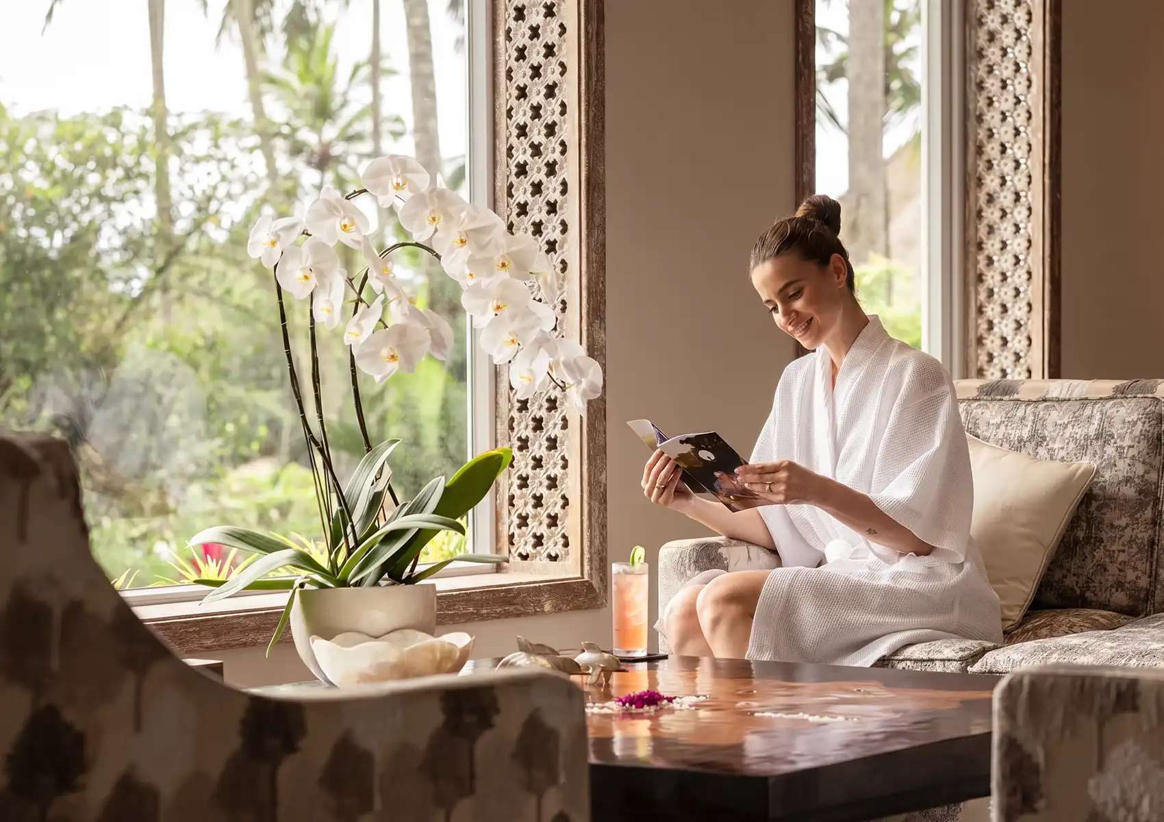 spa-in-bali A smiling woman in a white bathrobe relaxes with a brochure in a brightly lit seating area, sitting next to a potted white orchid and a large window overlooking lush tropical greenery, as she prepares for her reflexology ubud session.