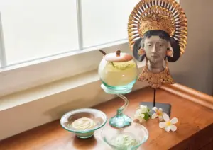Glass bowls and jars filled with natural body scrub powders and creams displayed on a wooden table beside a traditional Balinese carved statue and frangipani flowers at a spa in Ubud.