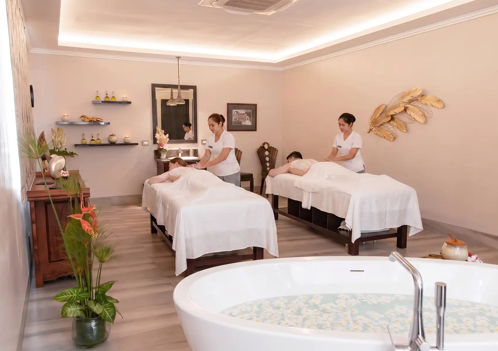 A couple receiving a relaxing treatment as therapists apply a nourishing Yoghurt body mask to their backs in a bright, luxurious spa room featuring a bathtub filled with flower petals.