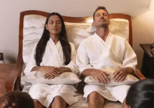 A relaxed couple wearing white bathrobes sit side-by-side with their eyes closed, enjoying a soothing treatment at a spa in Ubud.