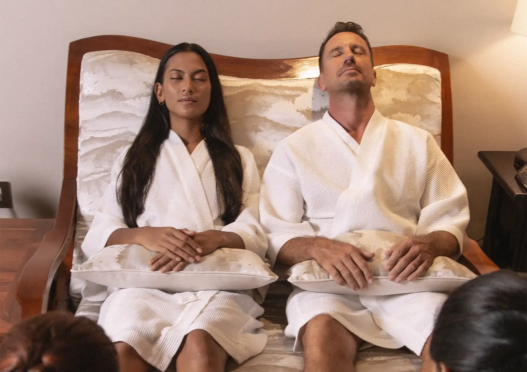A relaxed couple wearing white bathrobes sit side-by-side with their eyes closed, enjoying a soothing treatment at a spa in Ubud.