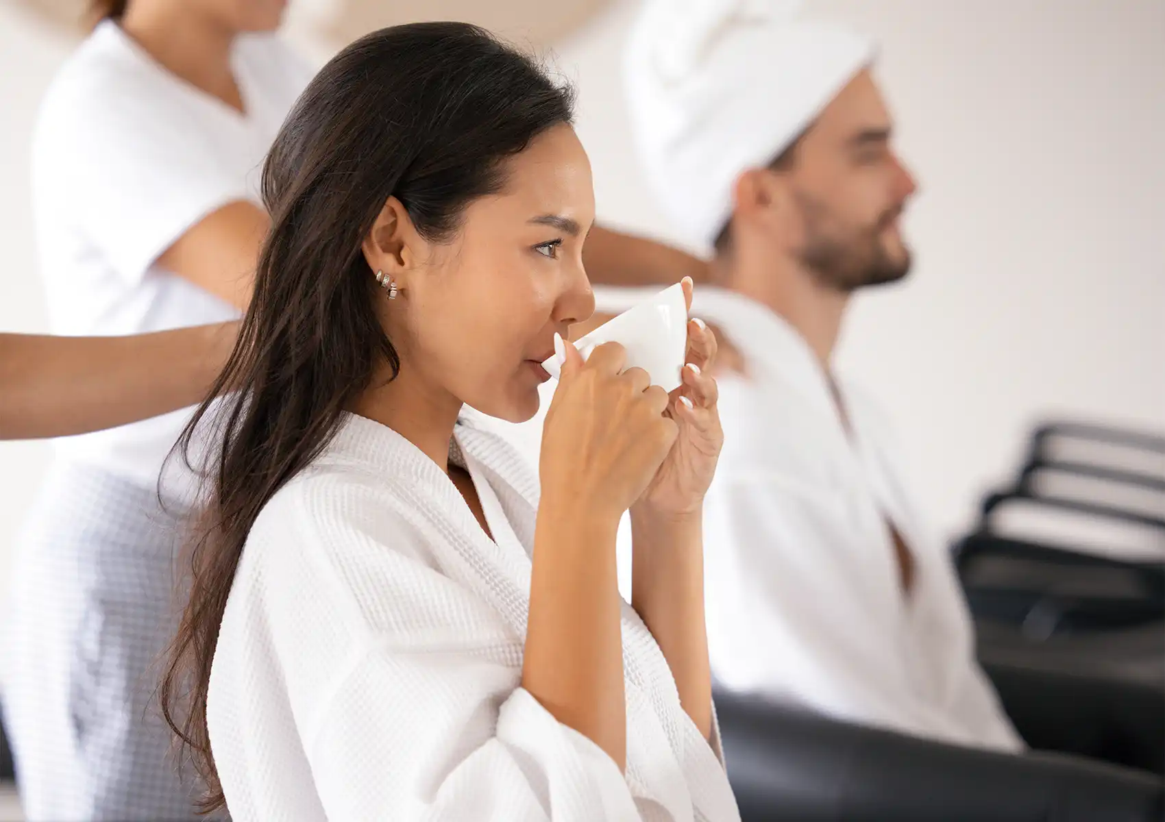 A relaxed woman in a white bathrobe smiles and enjoys a warm drink while receiving a soothing neck and shoulder massage from a therapist during a luxury hair treatment Ubud experience, with another spa guest sitting in the background.