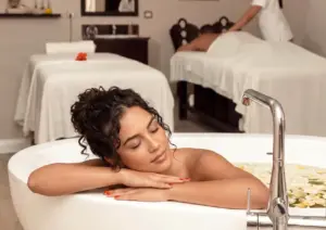 A serene woman with her eyes closed resting on the edge of a white bathtub during a soothing relaxation bath Ubud, with another guest receiving a massage in the tranquil spa background.