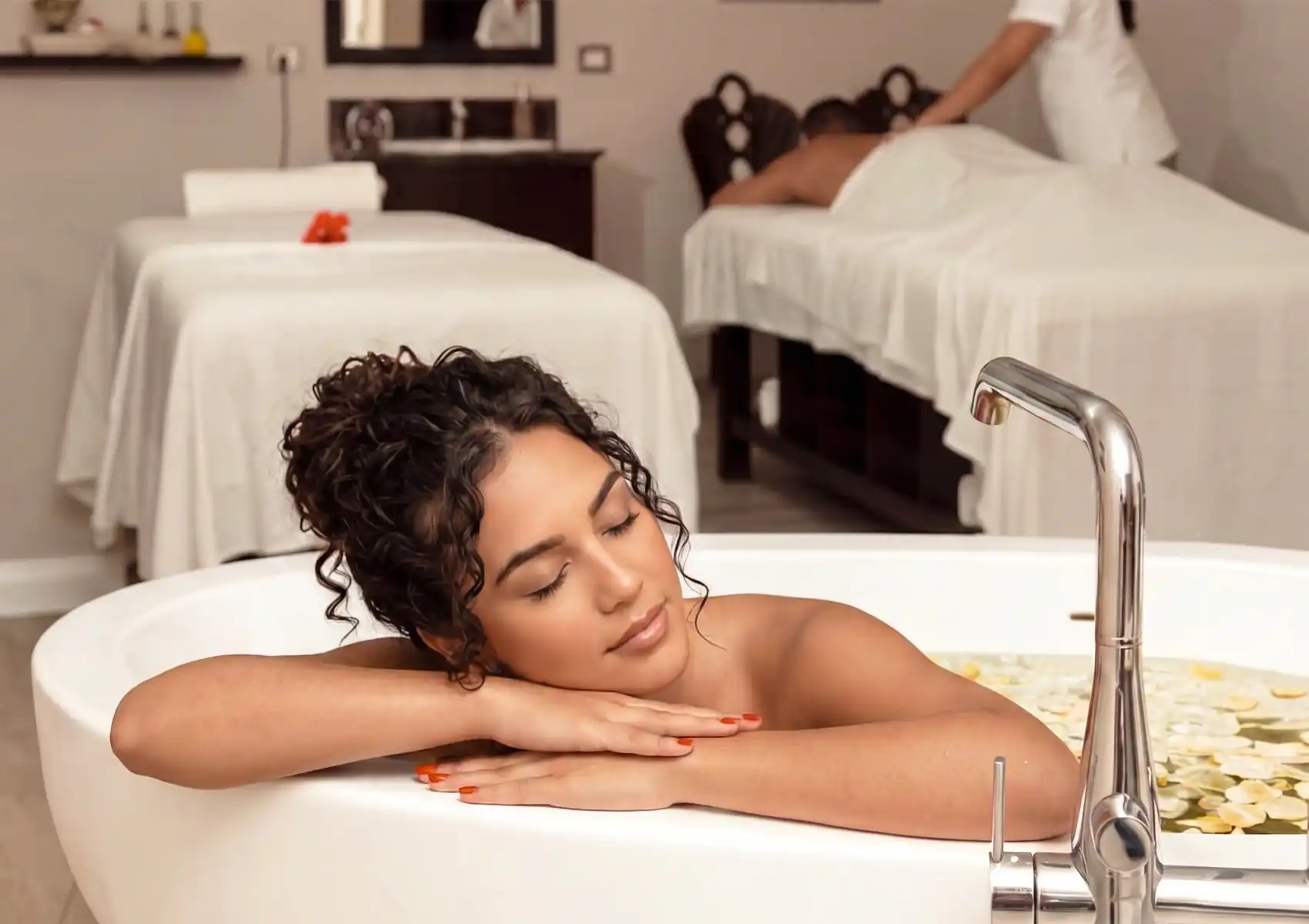 A serene woman with her eyes closed resting on the edge of a white bathtub during a soothing relaxation bath Ubud, with another guest receiving a massage in the tranquil spa background.