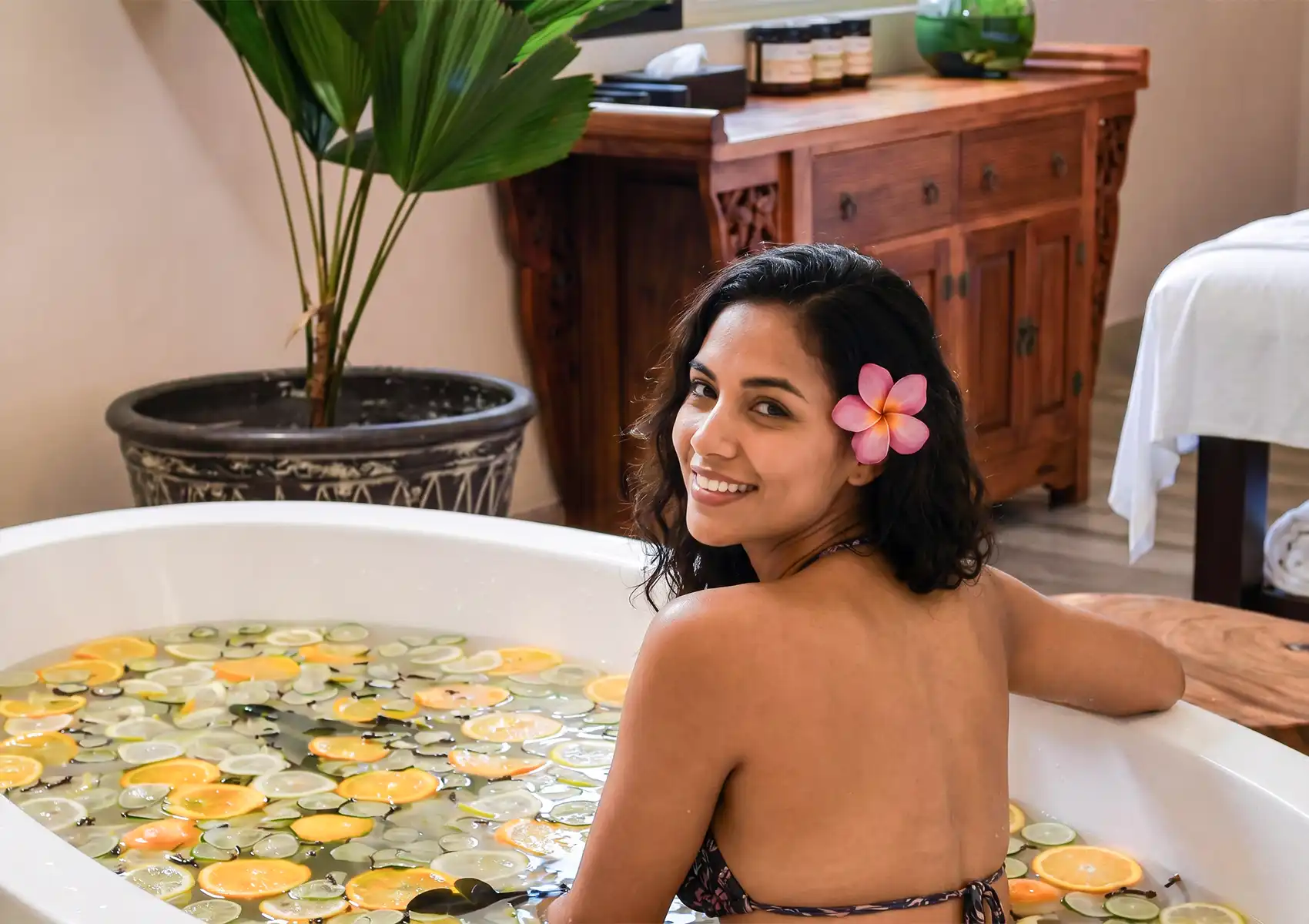 A smiling woman with a pink frangipani flower in her hair enjoying a refreshing citrus and spice-filled relaxation bath Ubud inside a beautiful tropical spa setting.