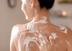 A close-up of a spa therapist's hand gently applying a thick, creamy white Yoghurt body mask to a woman's back and shoulders during a soothing wellness treatment.