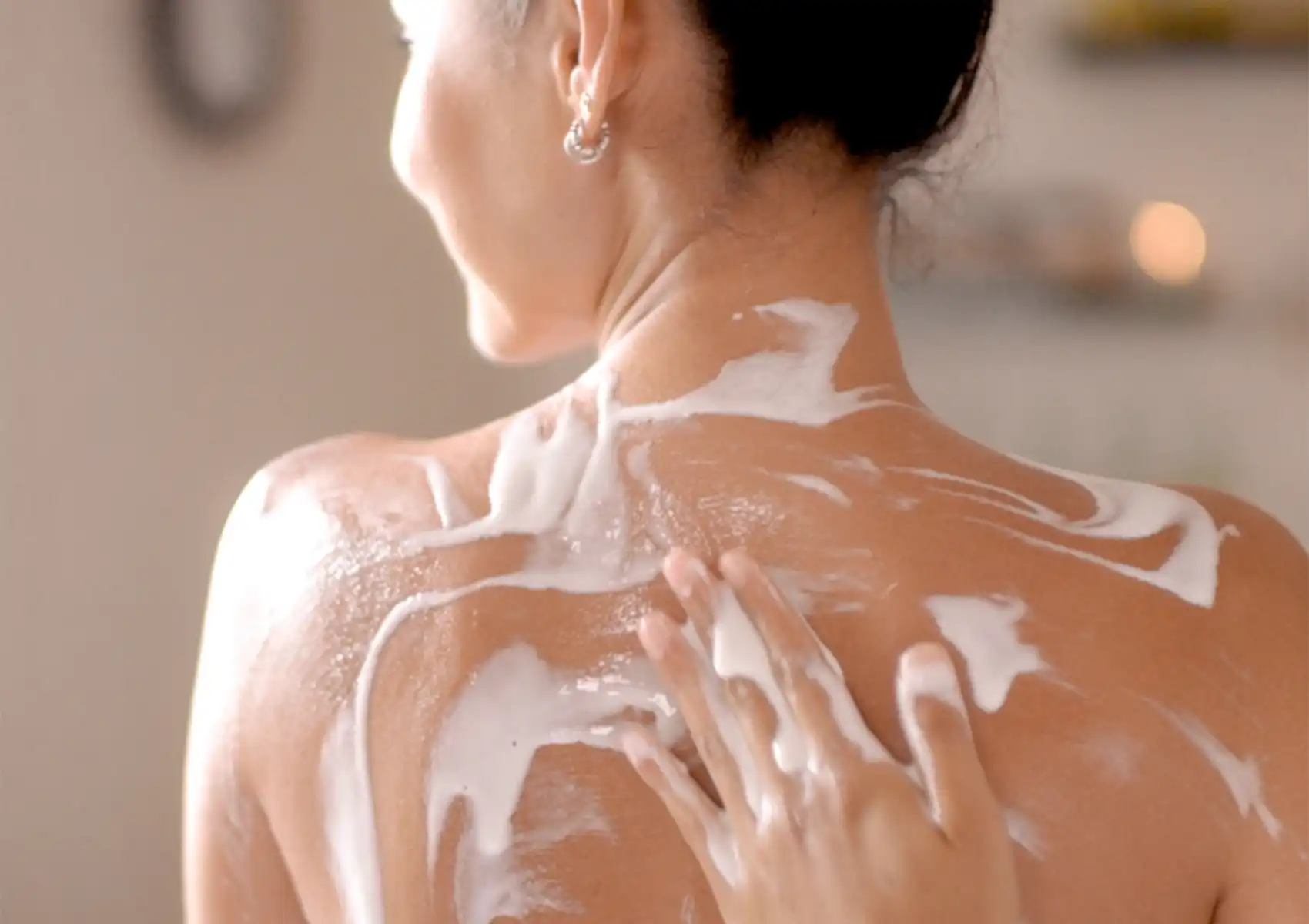 A close-up of a spa therapist's hand gently applying a thick, creamy white Yoghurt body mask to a woman's back and shoulders during a soothing wellness treatment.
