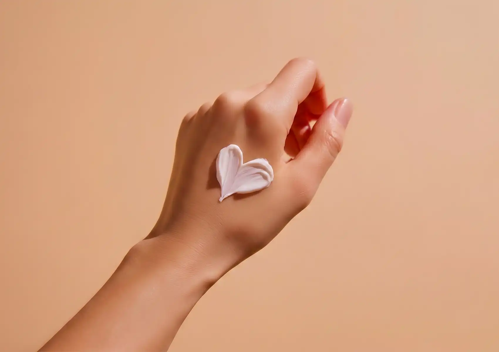 A close-up of a hand with a dollop of creamy white Yoghurt body mask shaped into a heart resting on the skin against a soft peach background.