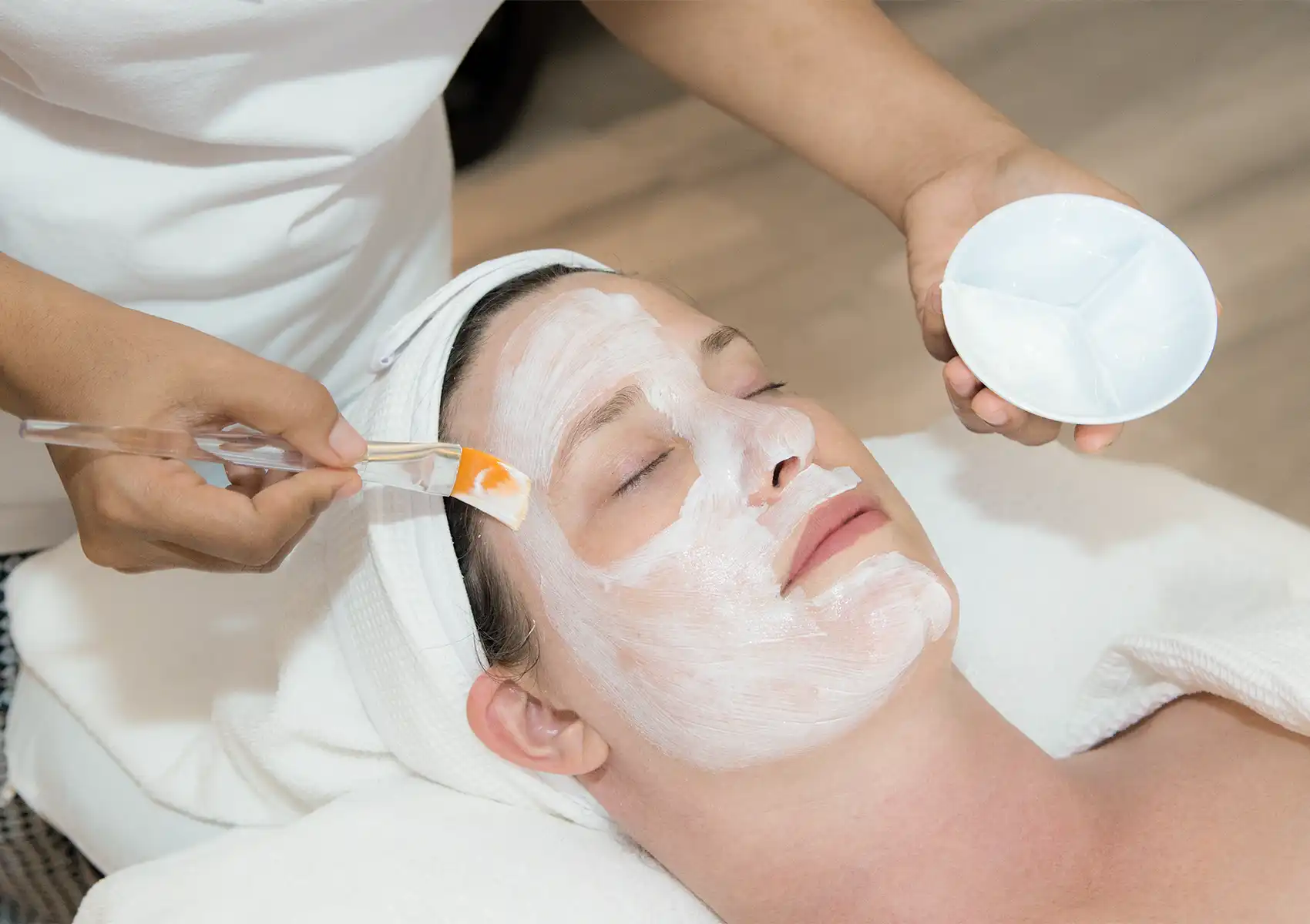 A professional spa therapist uses a brush to carefully apply a white facial mask to a relaxed woman at the best spa in ubud.