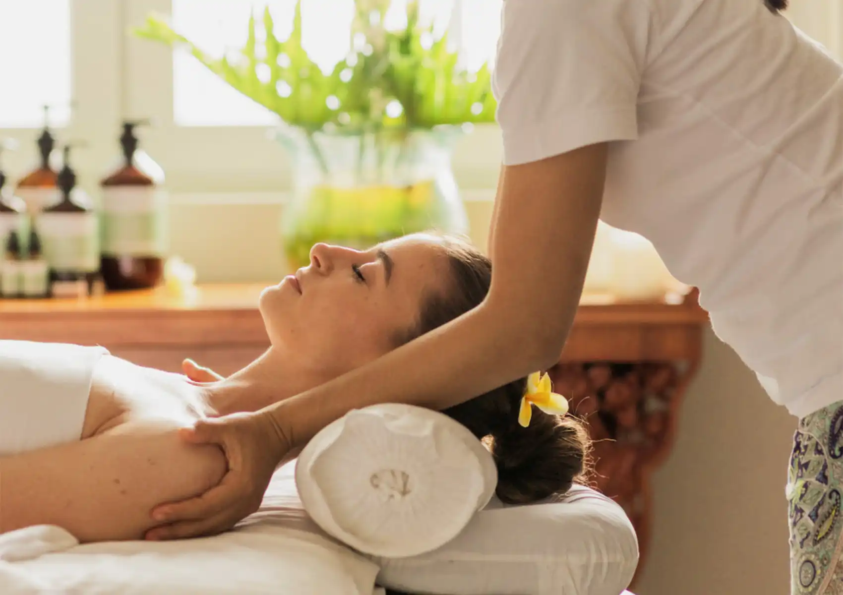 A woman with a frangipani flower in her hair receives a soothing shoulder and neck massage from a professional therapist in a bright, tranquil treatment room at the best spa in Ubud.