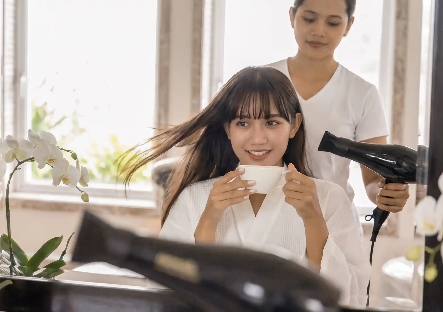A smiling woman in a white robe enjoying a warm drink while a stylist blow-dries her hair at the best spa in ubud.